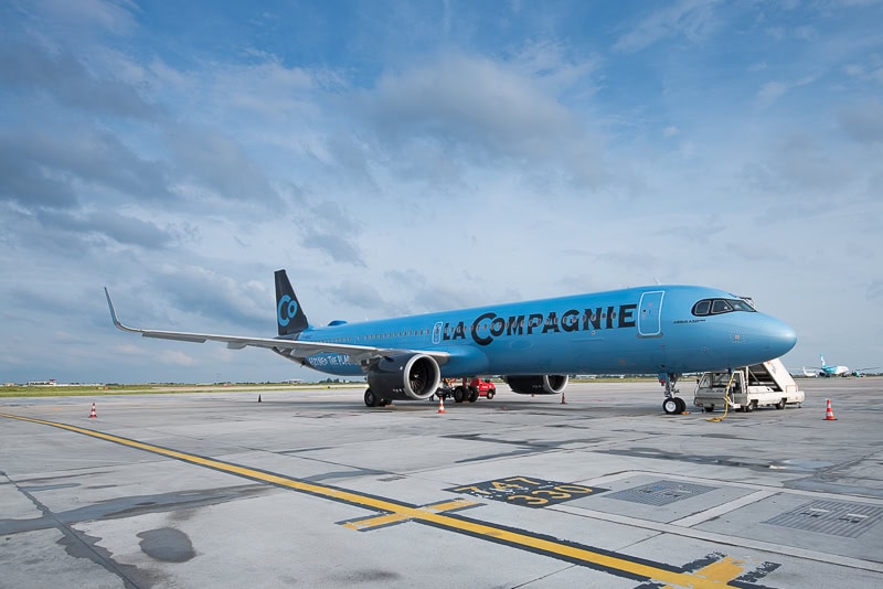 Ein hellblau lackiertes Flugzeug vom Typ Airbus A321neo der Fluggesellschaft La Compagnie steht auf einem Rollfeld unter einem bewölkten Tageshimmel.