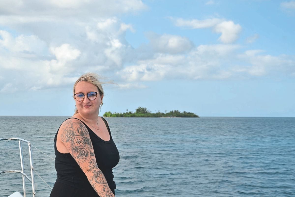 Eine Frau mit Brille und tätowiertem Arm steht an Deck eines Bootes vor dem Hintergrund des offenen Meeres und einer kleinen Insel bei bewölktem Himmel.