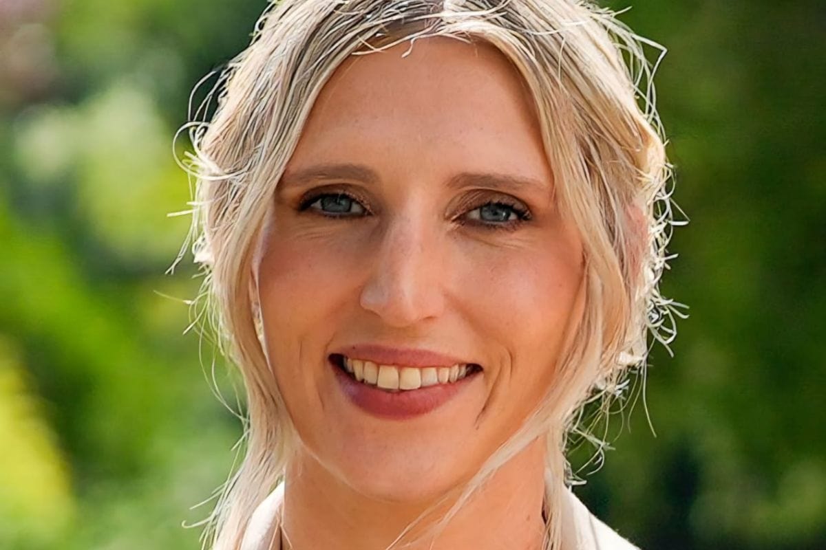 Nahaufnahme einer lächelnden Frau mit blondem Haar und blauen Augen vor einem unscharfen, grünen Hintergrund im Freien bei natürlichem Tageslicht aufgenommen.