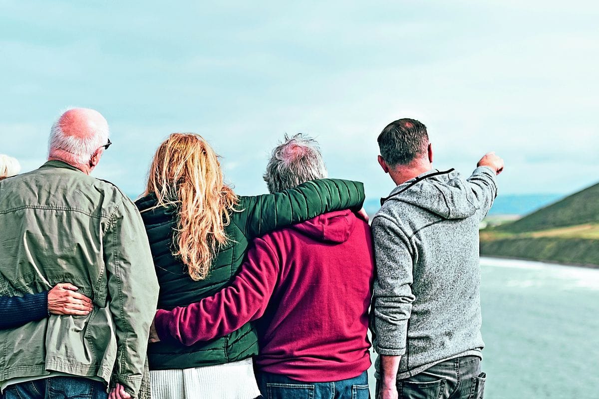 Vier Personen stehen in einer Reihe von hinten gesehen vor einer Küstenlandschaft, wobei eine Person mit dem Arm in die Ferne auf das Meer hinaus zeigt.