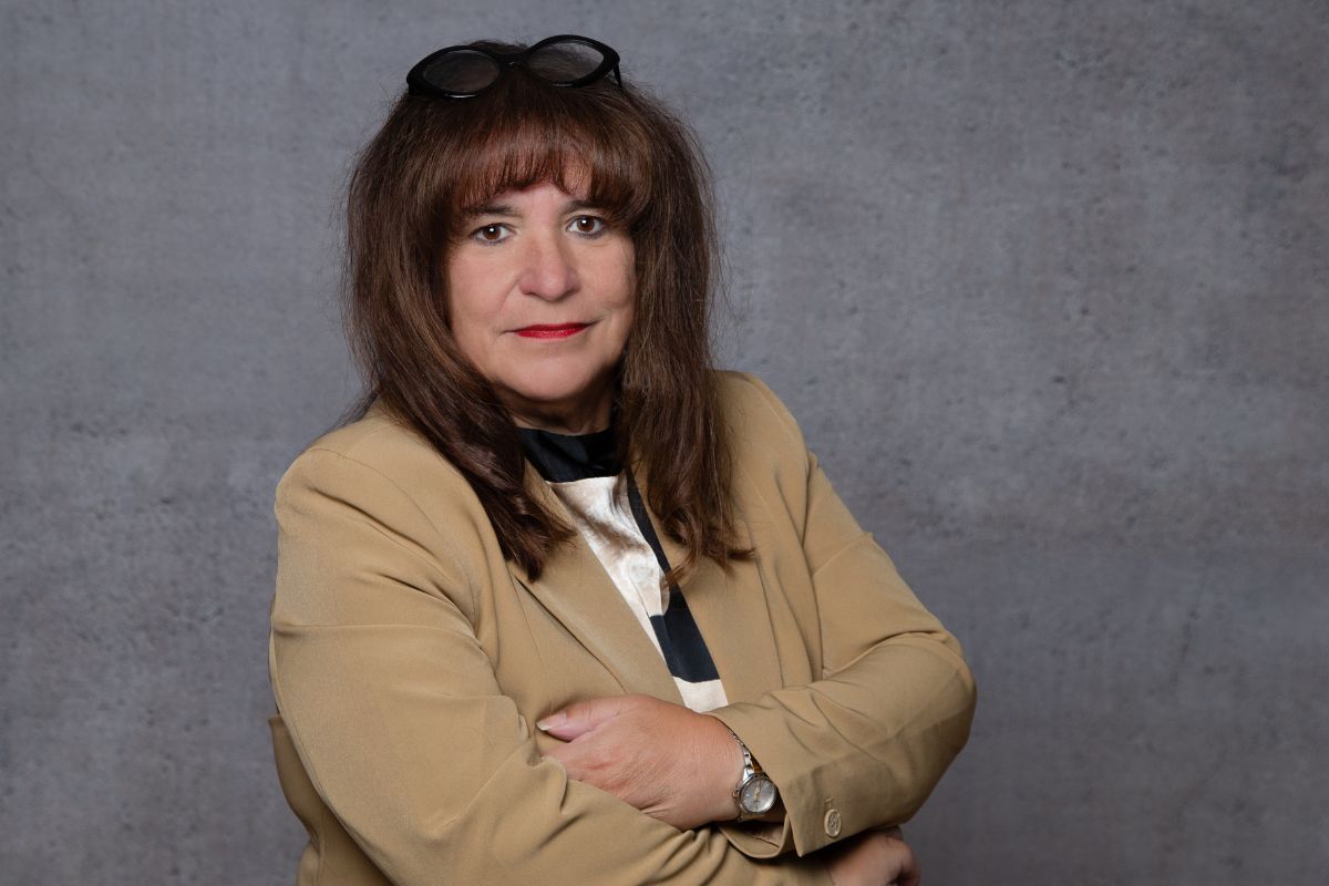 Eine Frau mit braunem Haar und Brille auf dem Kopf trägt einen beigen Blazer über einer gemusterten Bluse und steht vor einer grauen Betonwand im Porträt.