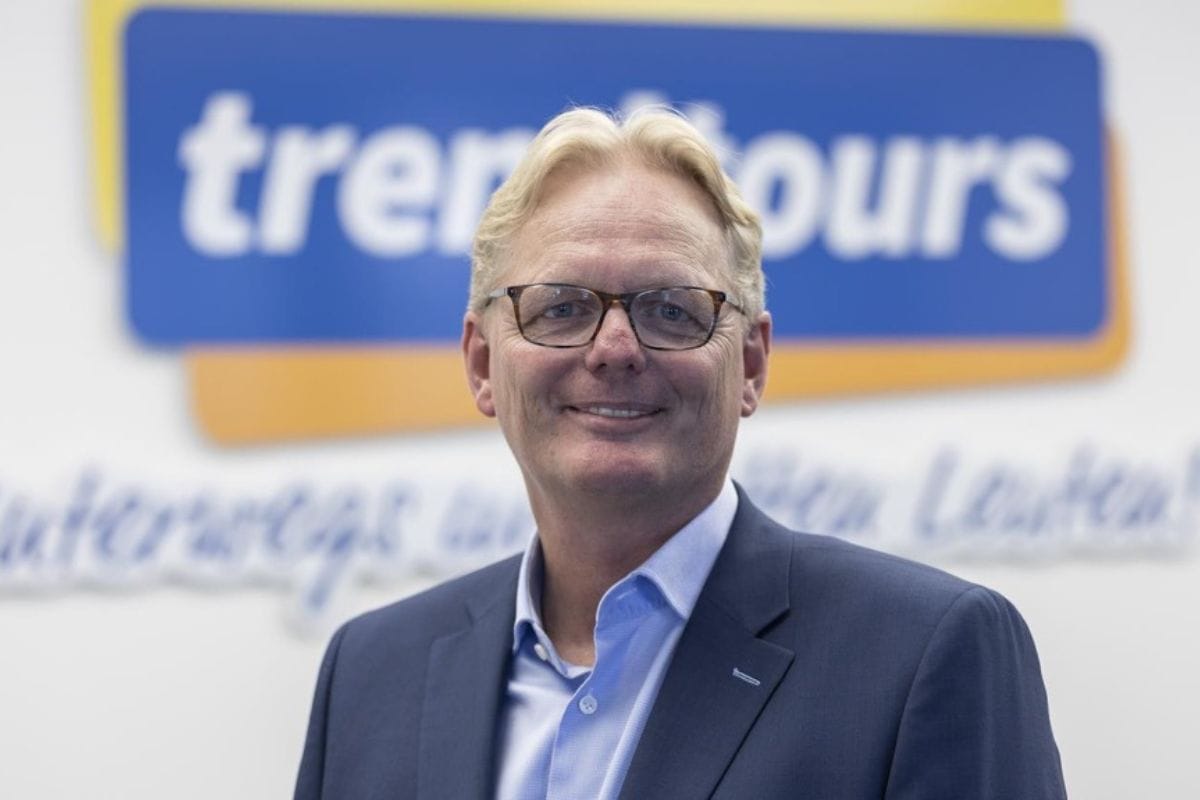 Ein hellhaariger Mann mit Brille trägt ein hellblaues Hemd und ein dunkelblaues Sakko. Im Hintergrund ist das Logo von trendtours an einer weißen Wand.