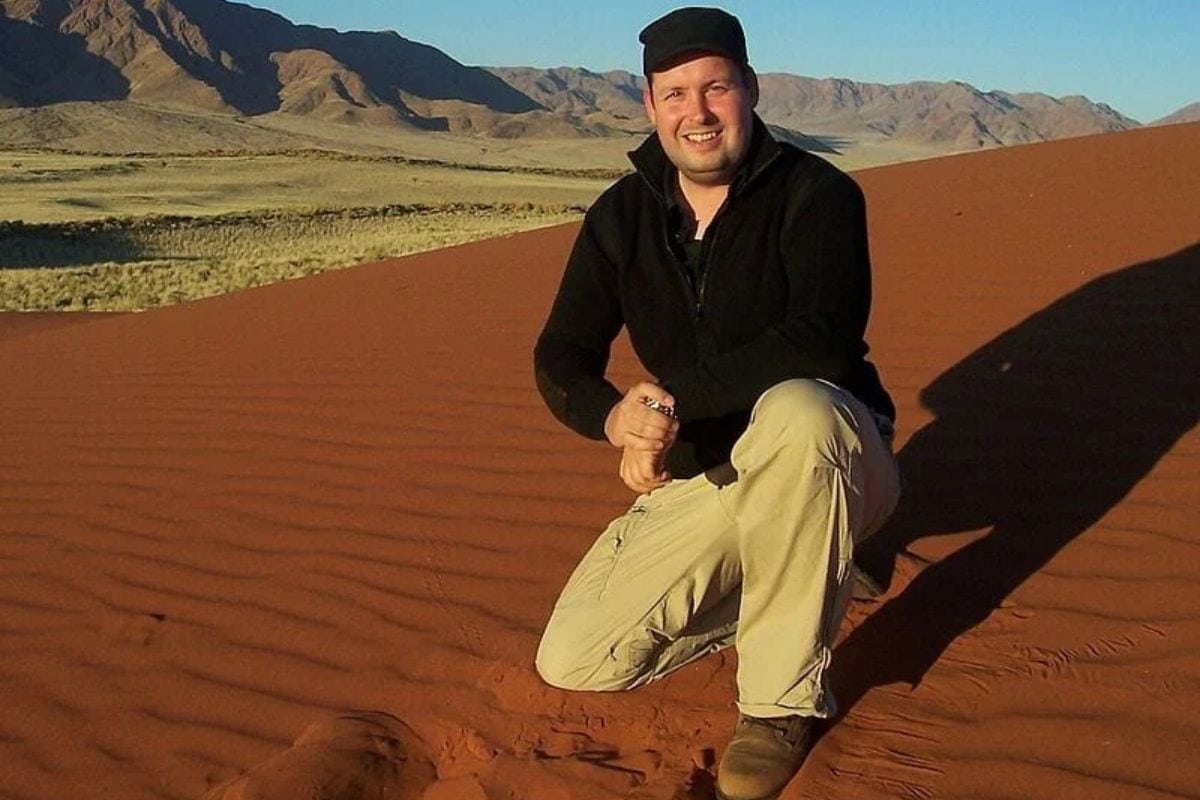 Ein Mann mit dunkler Jacke und Schirmmütze kniet auf einer rötlichen Sanddüne in einer weitläufigen Wüstenlandschaft mit Bergen im Hintergrund bei Tageslicht.