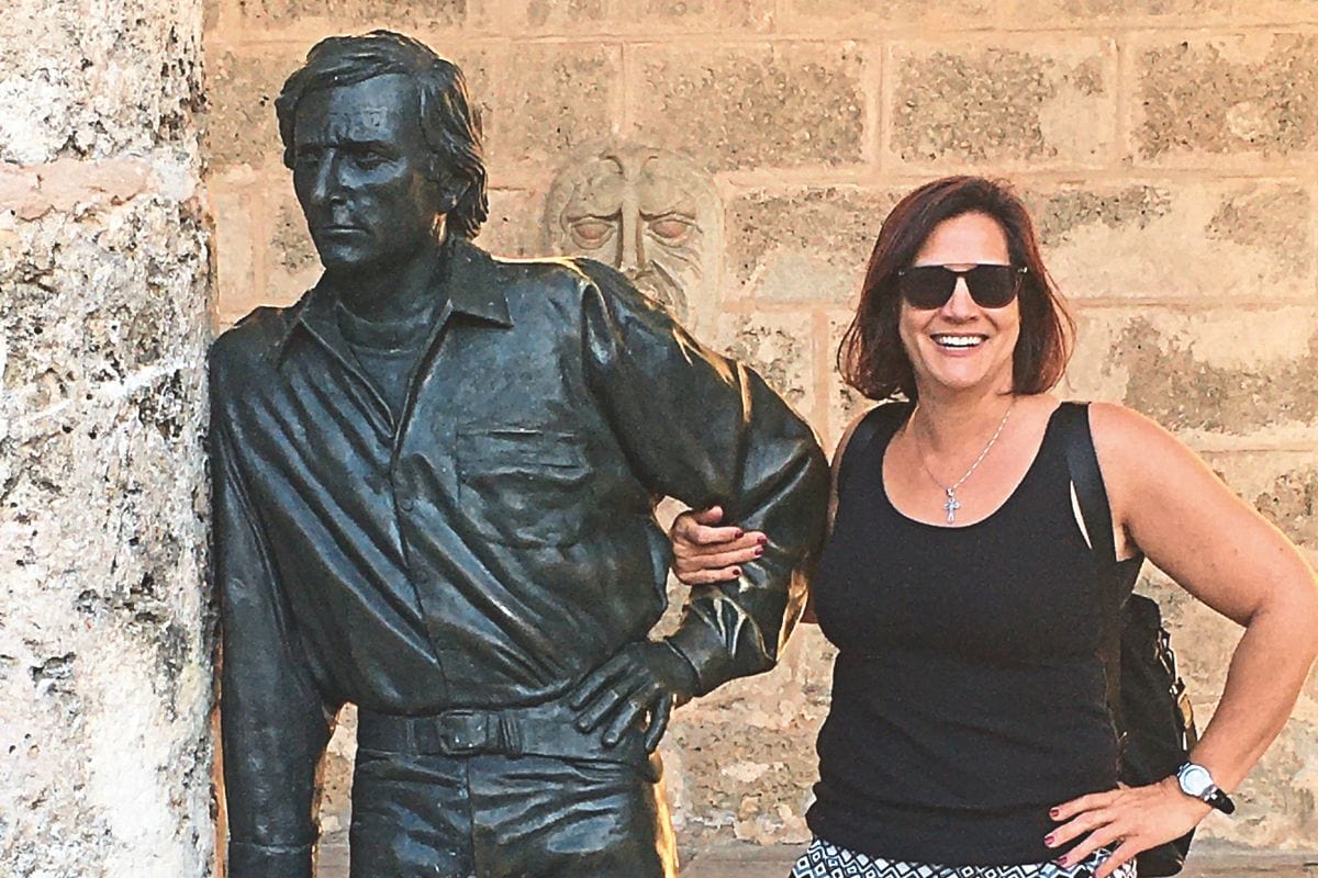 Eine lächelnde Frau mit Sonnenbrille steht neben einer lebensgroßen Bronzestatue eines Mannes vor einer rustikalen Steinmauer in einem Außenbereich.