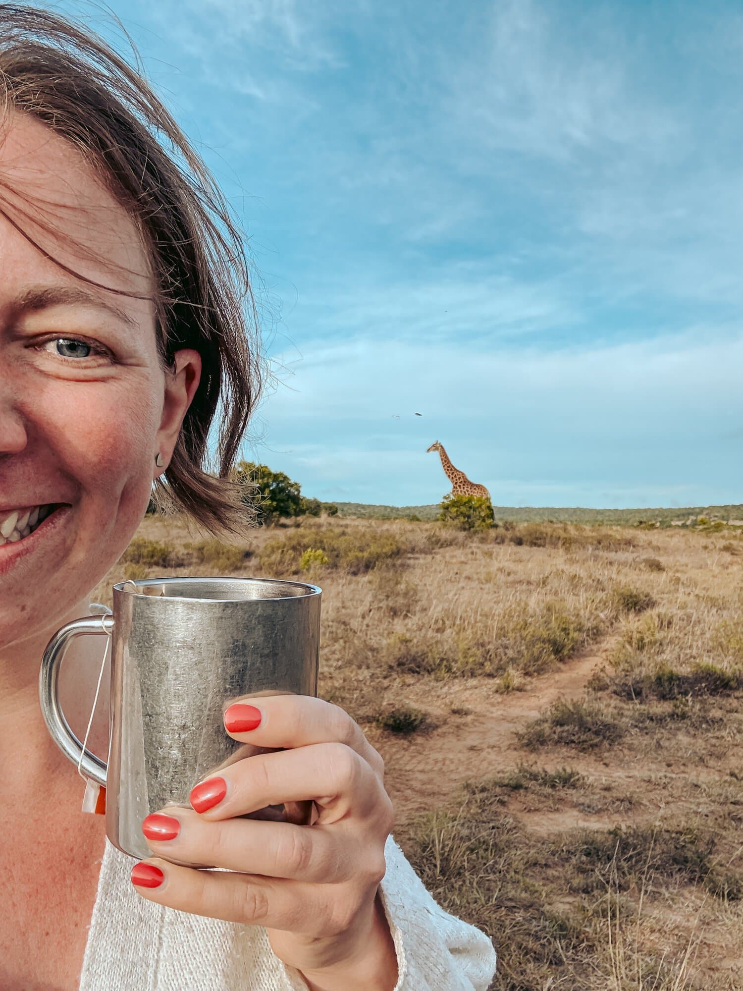 Eine Frau hält einen silbernen Metallbecher in der Hand, während im Hintergrund eine Giraffe in einer weiten, trockenen Savannenlandschaft zu sehen ist.