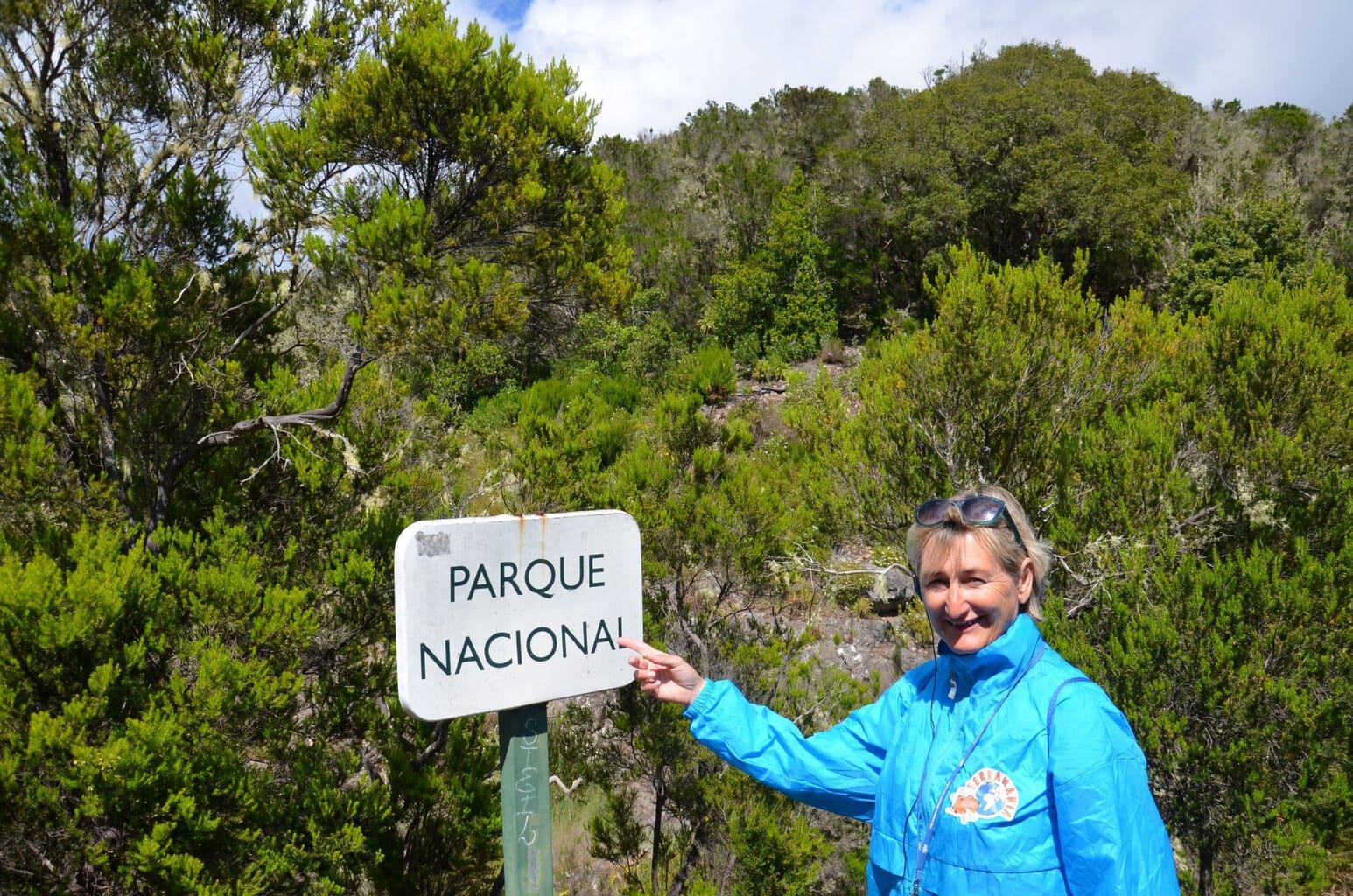 Eine Frau in einer blauen Jacke steht vor einem Schild mit der Aufschrift Parque Nacional und zeigt mit ihrem Finger auf den Text vor einer Waldkulisse.