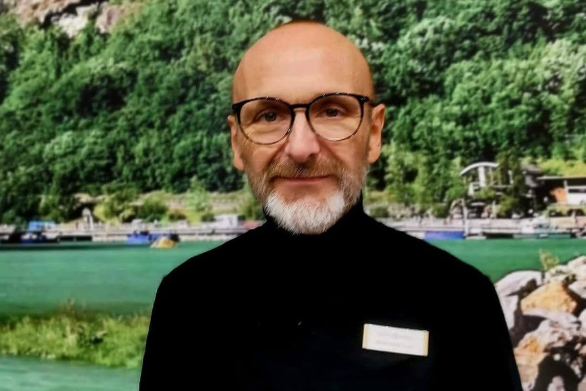 Ein Mann mit Glatze, Brille und grauem Bart trägt einen schwarzen Rollkragenpullover vor einem Hintergrundbild mit einer grünen Seen- und Berglandschaft.