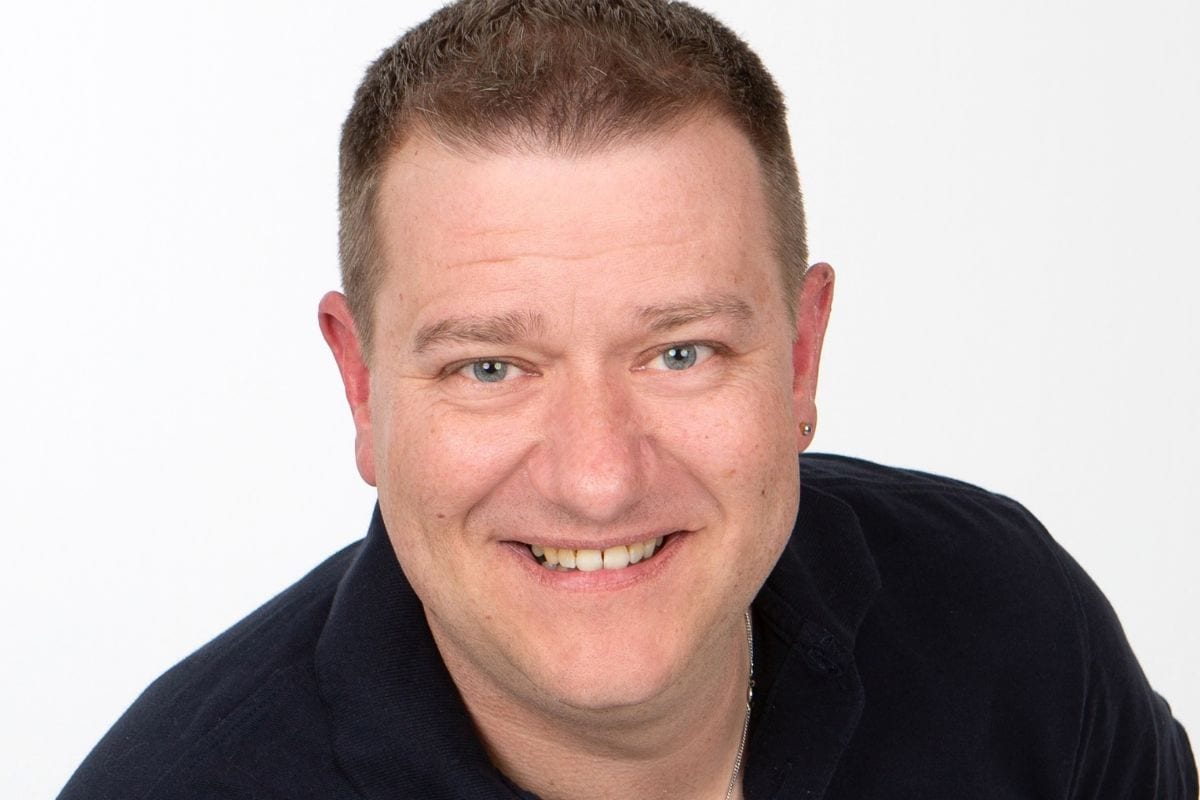 Ein Mann mit kurzem braunem Haar und blau-grauen Augen lächelt freundlich in die Kamera. Er trägt ein dunkelblaues Poloshirt vor einem weißen Hintergrund.