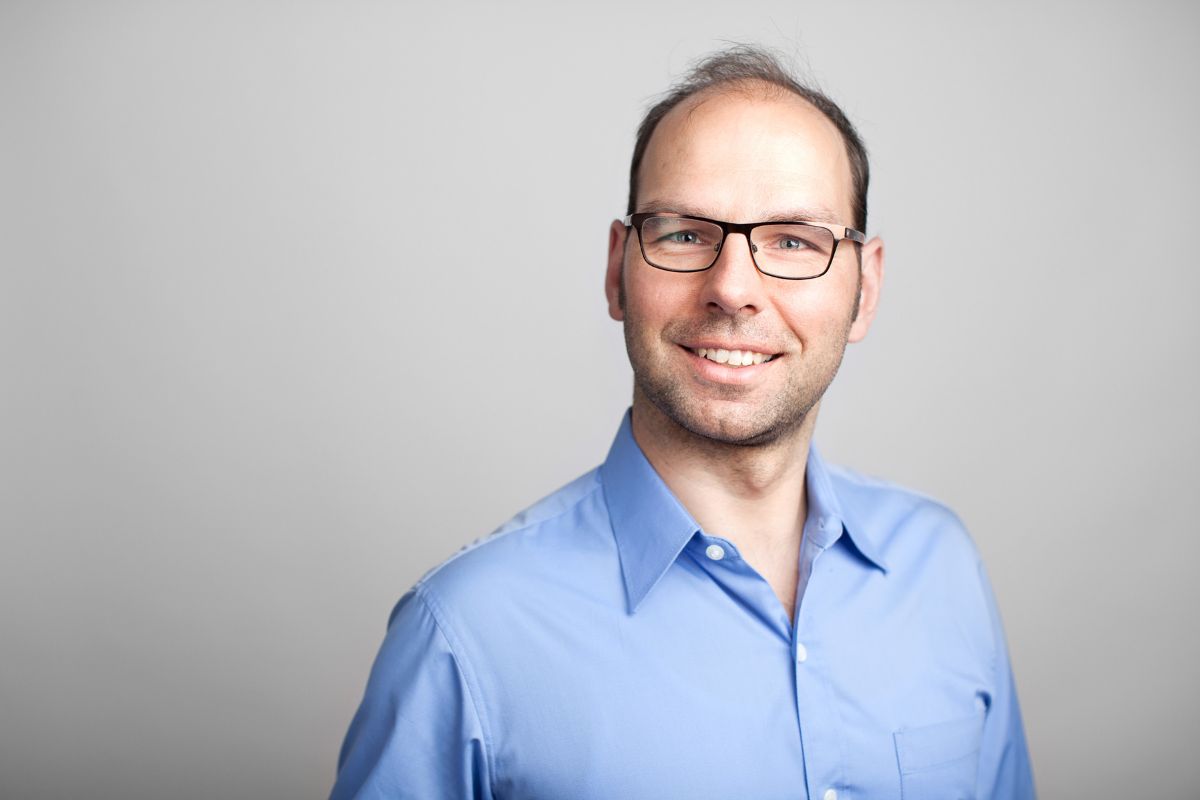 Ein Mann mit kurzem Haar und Brille trägt ein hellblaues Hemd und lächelt freundlich in die Kamera vor einem neutralen, hellgrauen Hintergrund im Studio.