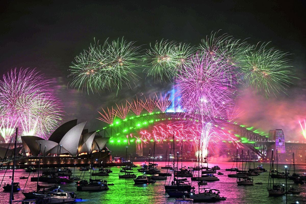 Ein nächtliches Feuerwerk in Grün und Pink über der beleuchteten Sydney Harbour Bridge und dem Opernhaus, mit zahlreichen Booten im dunklen Hafenwasser.