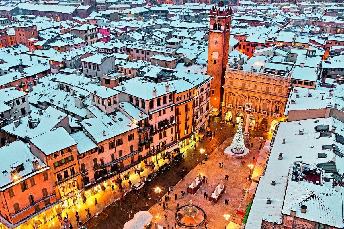 Luftaufnahme einer verschneiten italienischen Stadt mit einem beleuchteten Weihnachtsbaum auf einem zentralen Platz und einem historischen Backsteinturm.