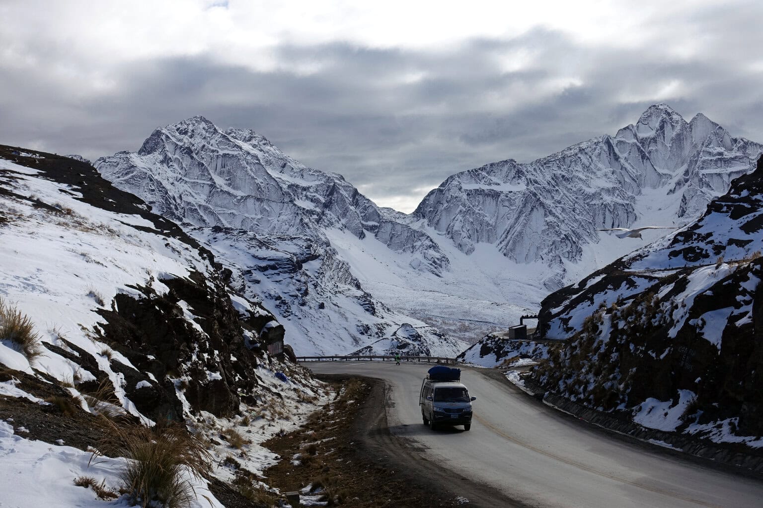 Ein Kleinbus fährt auf einer kurvigen Asphaltstraße durch eine schneebedeckte Gebirgslandschaft mit hohen, schroffen Gipfeln unter einem bewölkten Himmel.
