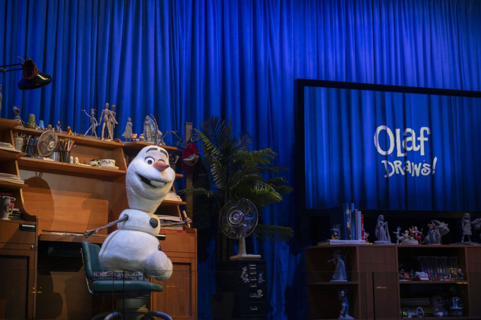 Ein freundlicher Schneemann sitzt auf einem Stuhl vor einem Schreibtisch mit verschiedenen Kunstmaterialien und einer großen blauen Vorhangkulisse.