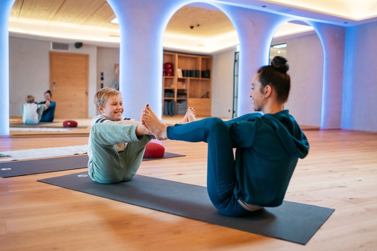 Eine Frau und ein Junge sitzen sich in einem hellen Fitnessraum auf Yogamatten gegenüber und berühren sich mit den Fußsohlen bei einer Dehnübung.