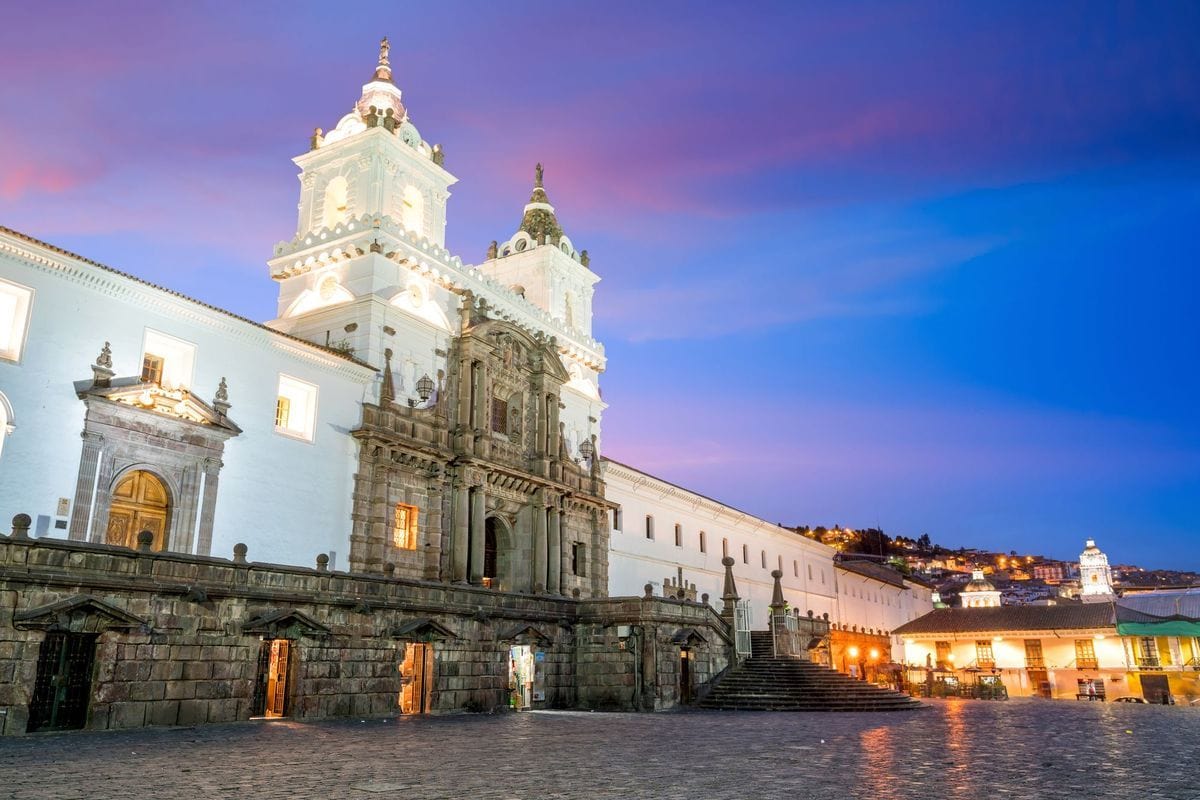 Die beleuchtete Fassade der Kirche San Francisco in Quito bei Dämmerung mit einem gepflasterten Vorplatz und einem violett-blauen Abendhimmel im Hintergrund.