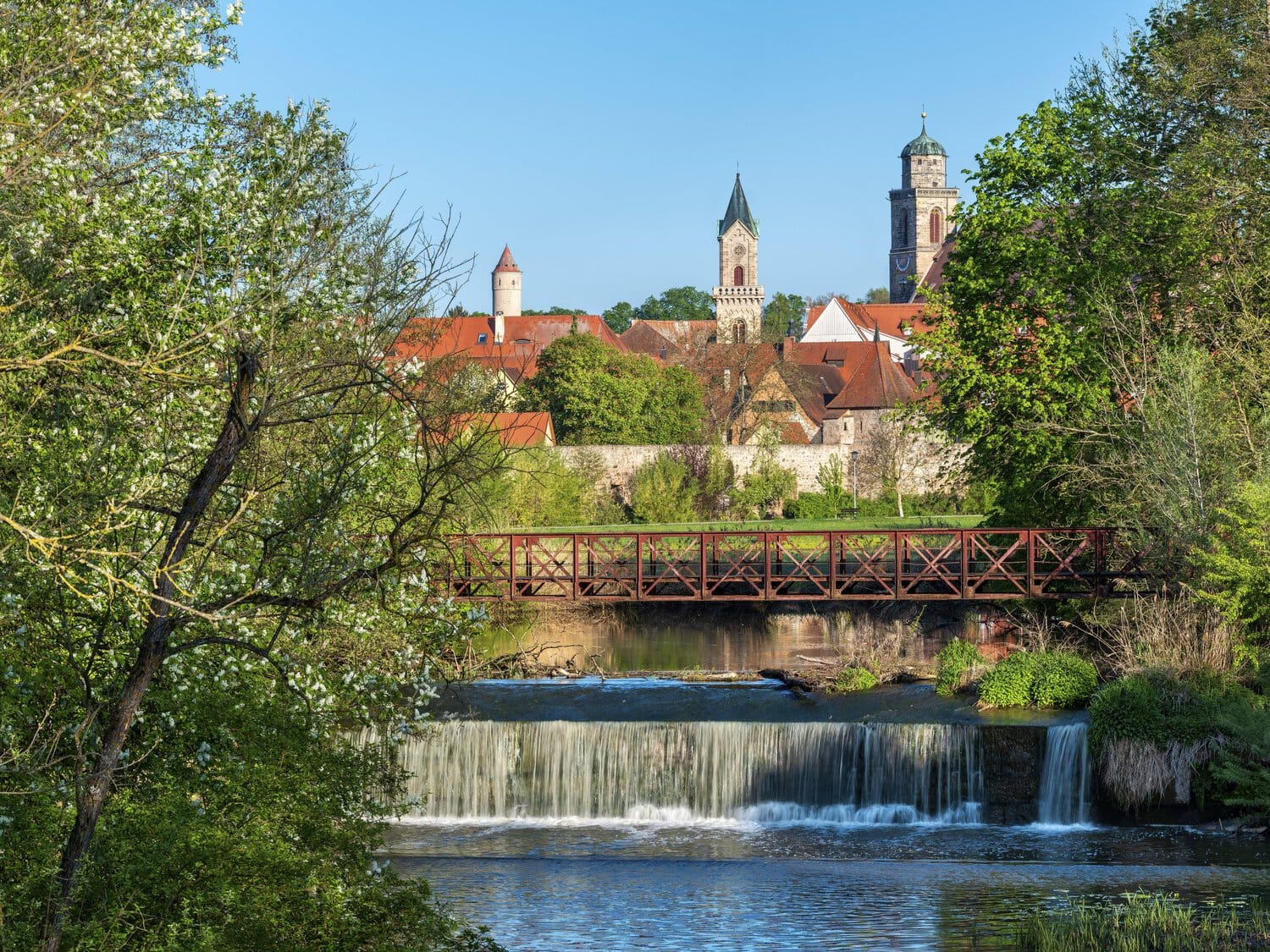 Blick auf eine historische Stadt mit Kirchtürmen und Stadtmauer hinter einem Fluss mit einem kleinen Wehr und einer metallenen Fußgängerbrücke im Vordergrund.