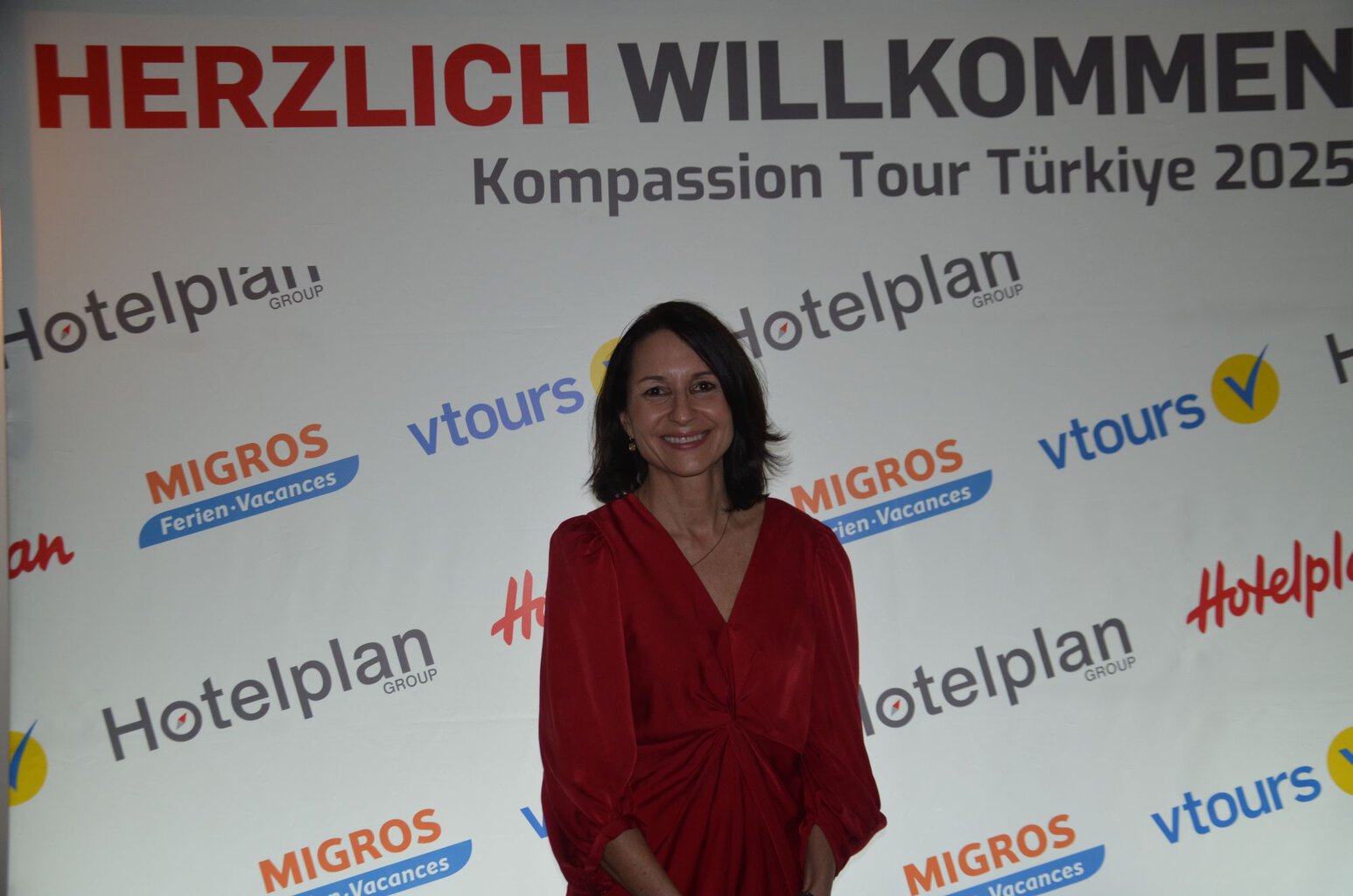 Eine lächelnde Frau mit schulterlangem dunklem Haar trägt ein rotes Kleid und steht vor einer Wand mit verschiedenen Firmenlogos und einer Begrüßung.
