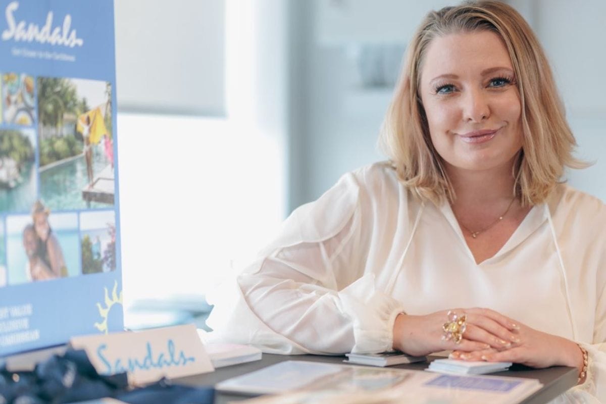 Eine blonde Frau mit weißer Bluse sitzt an einem Tisch mit Sandals-Werbematerialien und blickt freundlich lächelnd direkt in die Kamera des Betrachters.