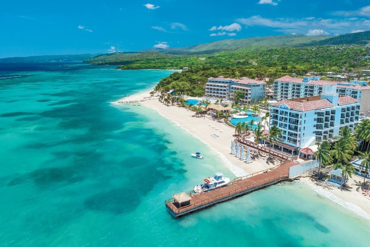 Luftaufnahme eines Resorts mit weißen Hotelgebäuden, einem langen Holzsteg, türkisfarbenem Meer und einem Sandstrand vor einer grünen Hügellandschaft.