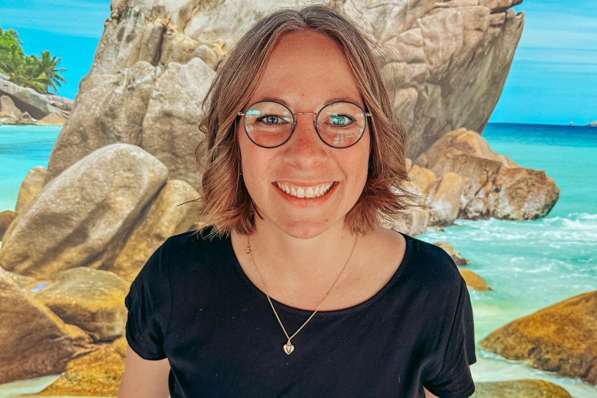 Eine junge Frau mit halblangem braunem Haar und einer runden Brille lächelt vor einer Fototapete mit einer tropischen Strandkulisse und großen Felsen.