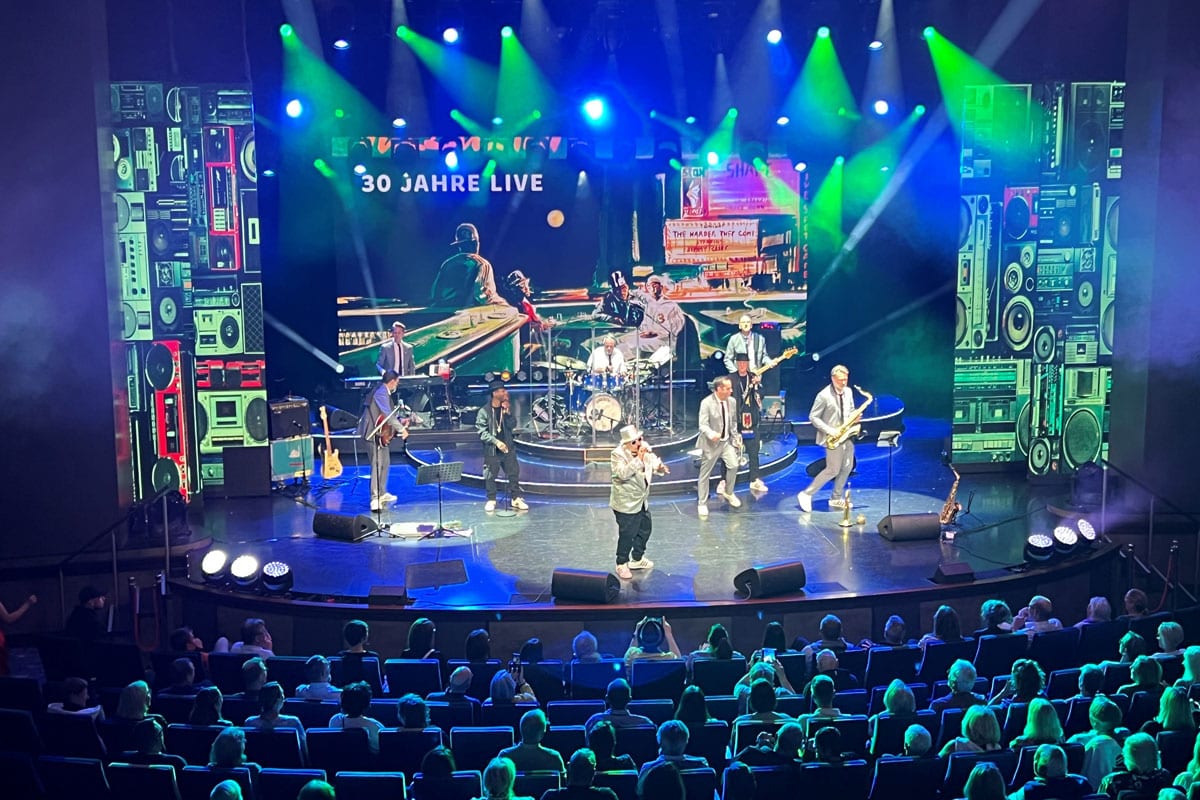 Eine Musikband tritt auf einer beleuchteten Bühne vor einem Publikum auf, im Hintergrund ist eine große Leinwand mit der Aufschrift 30 JAHRE LIVE zu sehen.