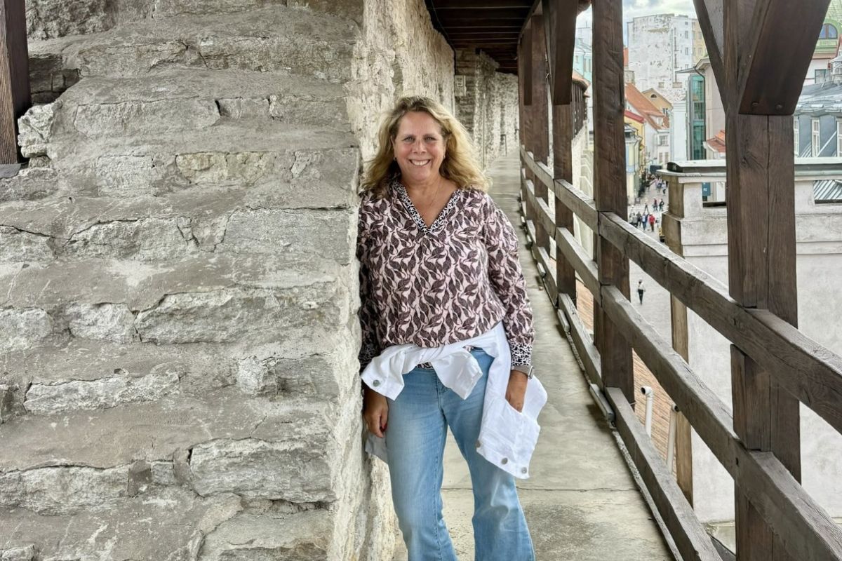 Eine blonde Frau mit gemusterter Bluse und Jeans steht lächelnd auf einem überdachten Wehrgang aus Stein neben einer historischen Stadtmauer in Tallinn.