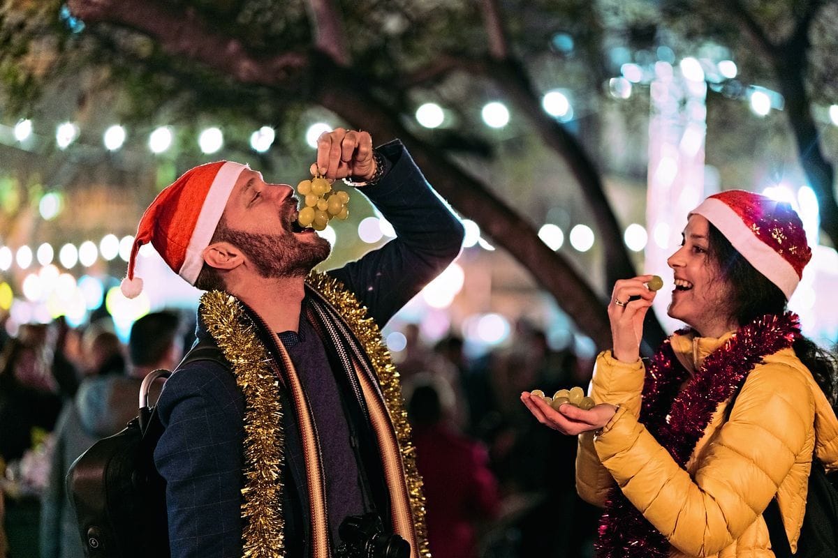 Ein Mann und eine Frau mit Weihnachtsmützen essen im Freien bei abendlicher Lichterkette Trauben aus der Hand vor einer unscharfen Menschenmenge im Hintergrund.