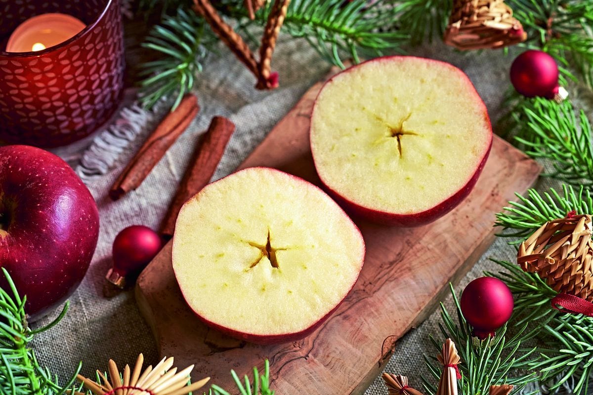 Zwei Apfelhälften liegen auf einem Holzbrett, umgeben von Tannenzweigen, Zimtstangen, kleinen roten Weihnachtskugeln und einem brennenden roten Windlicht.