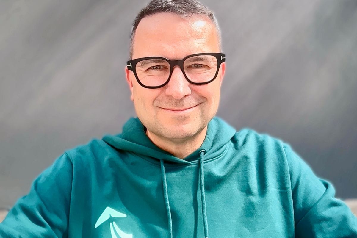 Ein Mann mit kurzem Haar und einer schwarzen Brille lächelt freundlich in die Kamera. Er trägt einen smaragdgrünen Kapuzenpullover vor einem grauen Hintergrund.