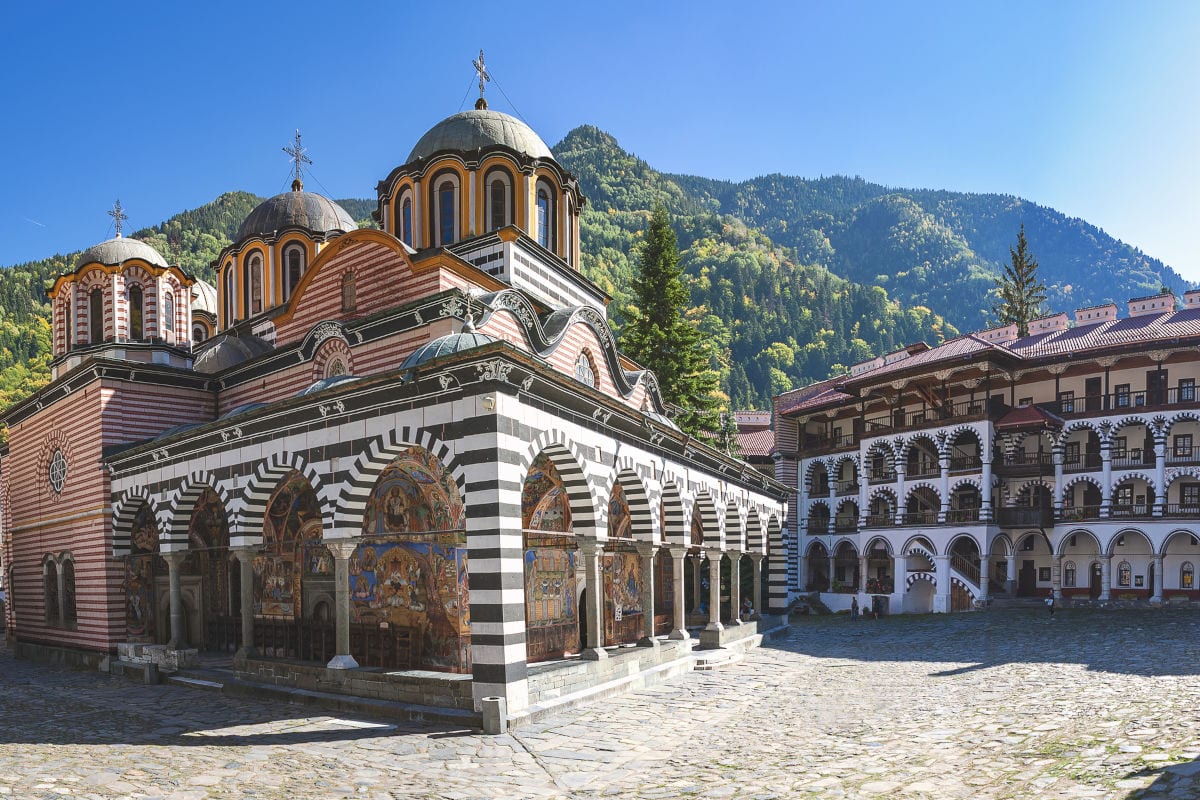 Das Rila-Kloster in Bulgarien mit seiner gestreiften Kirche, Arkadengängen und dem gepflasterten Innenhof vor einer bewaldeten Bergkulisse bei blauem Himmel.
