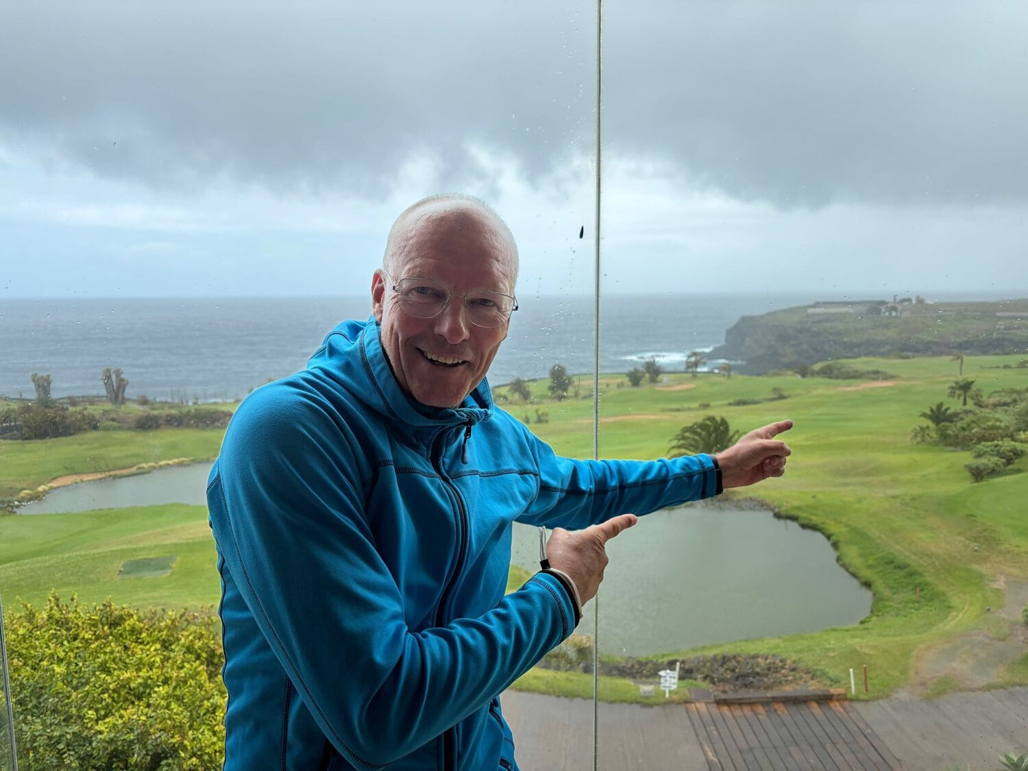 Ein lächelnder Mann mit Brille und blauer Fleecejacke steht vor einer großen Fensterscheibe und zeigt mit beiden Händen auf eine grüne Golfplatzanlage.