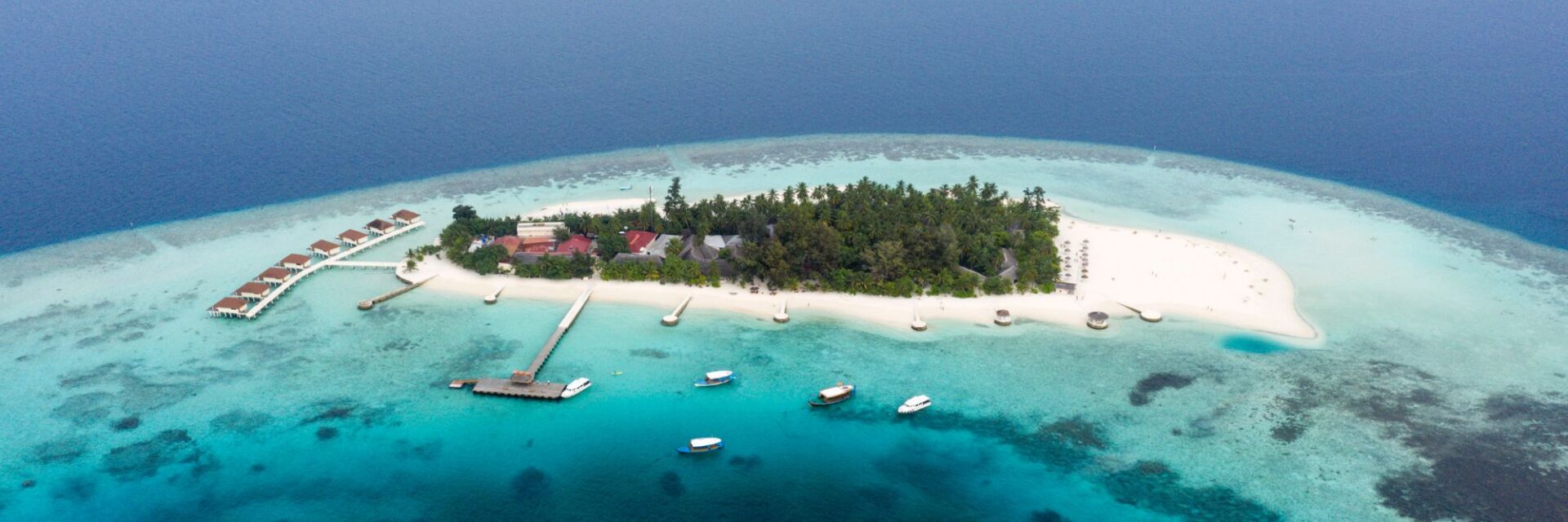 malediven-steuer Luftaufnahme einer tropischen Insel mit weißem Sandstrand, Palmen, einem langen Holzsteg und mehreren Booten im türkisfarbenen Wasser des Indischen Ozeans.
