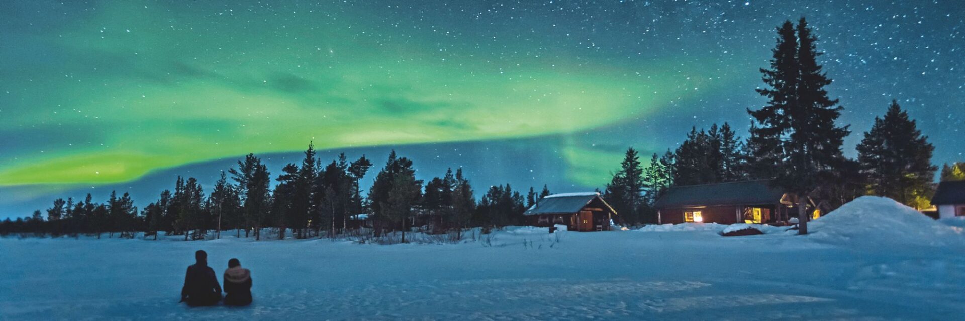 Zwei Personen sitzen in einer verschneiten Winterlandschaft vor Holzhütten und betrachten grüne Polarlichter am nächtlichen Sternenhimmel über dem Wald.