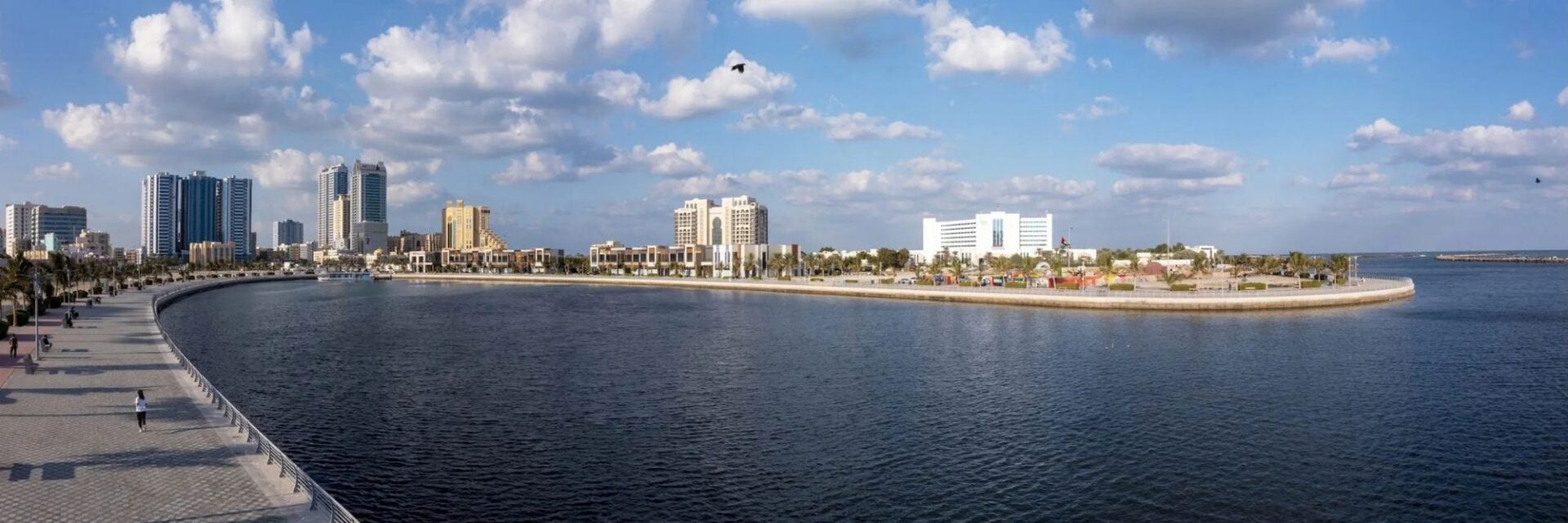 Website Header - 2026-09-23T113157.919 Panoramablick auf eine Uferpromenade mit gepflastertem Weg, angrenzendem Wasserbecken und einer modernen Skyline mit Hochhäusern unter blauem Sommerhimmel.