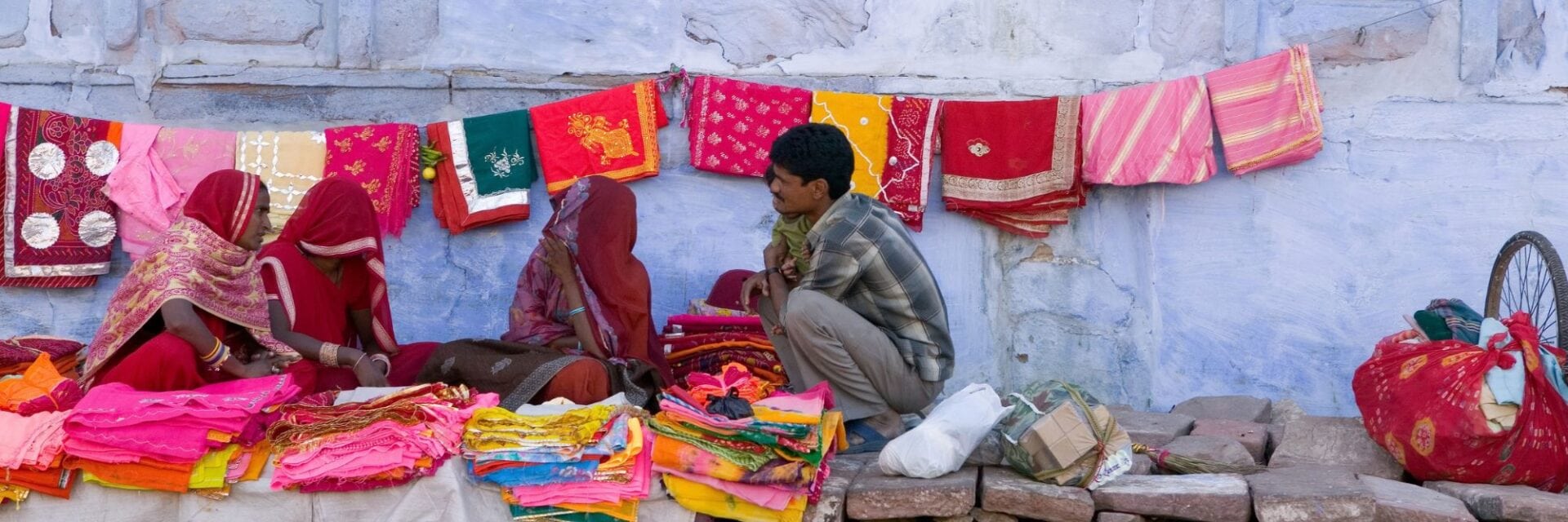 indien-jodhpur-markt Drei Frauen in traditioneller Kleidung und ein Mann sitzen vor einer hellblauen Mauer, an der bunte Stoffe und Tücher zum Verkauf aufgehängt sind.