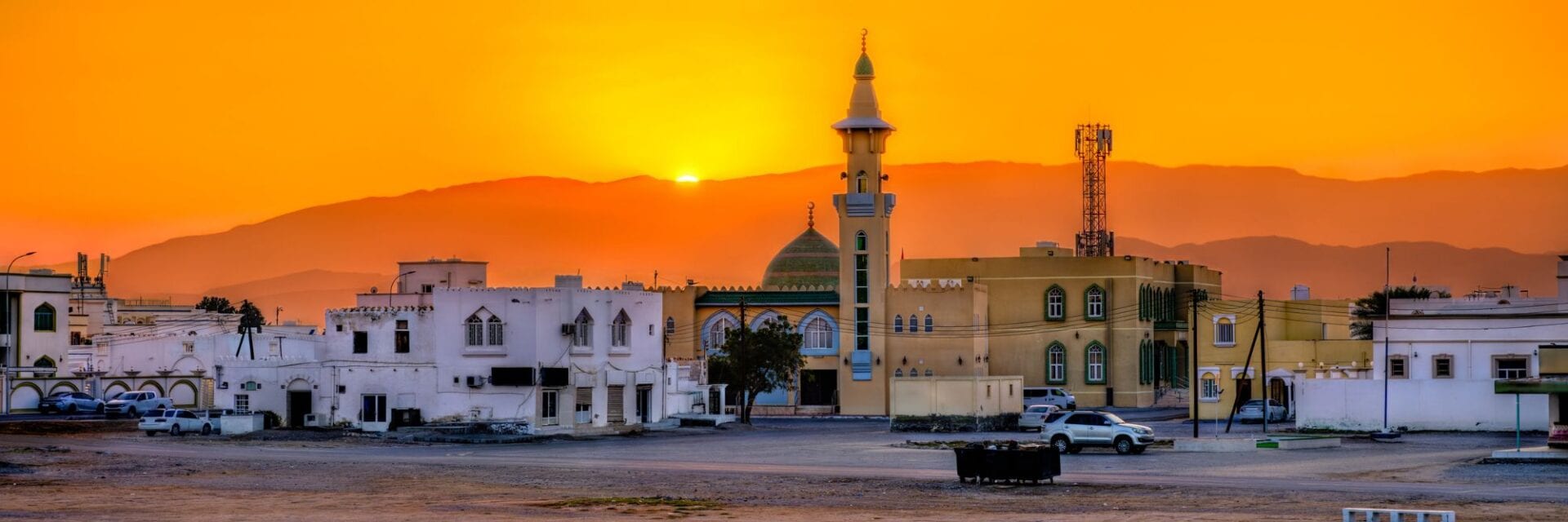 oman-sonnenuntergang Eine Moschee mit Minarett und Kuppel steht in einer Wüstenstadt vor einer Bergkette, während die Sonne in einem kräftigen Orange am Horizont untergeht.