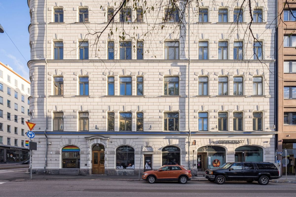 Die helle Fassade eines mehrstöckigen Wohn- und Geschäftshauses mit zahlreichen Fenstern, Erdgeschoss mit Ladenlokalen und parkenden Autos auf der Straße.