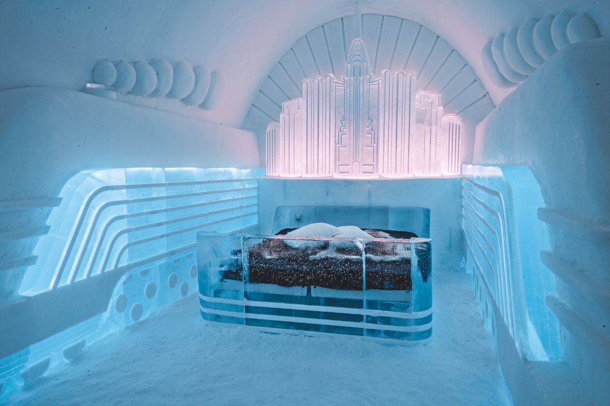 Ein Hotelzimmer aus Eis mit einem Bett, das von einer beleuchteten Skulptur im Stil eines Wolkenkratzers an der Wand und blauen Lichtakzenten dominiert wird.