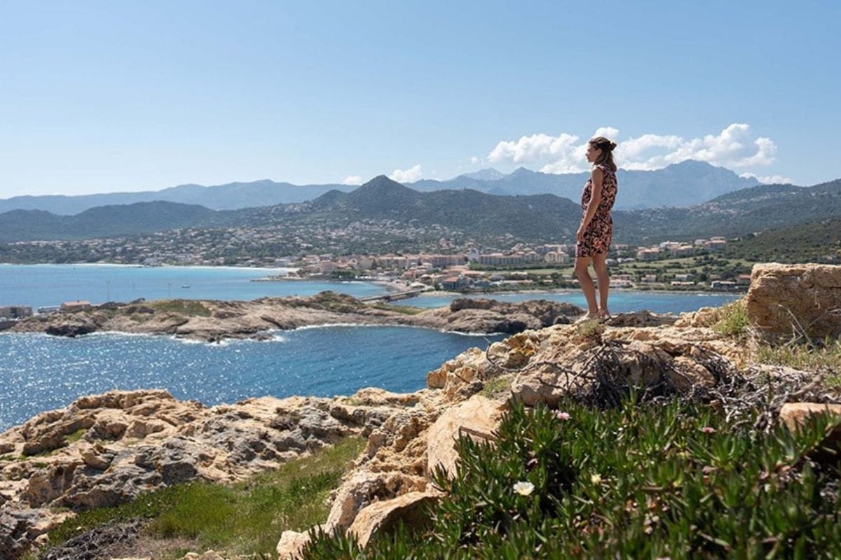 Eine Frau in einem gemusterten Kleid steht auf einem felsigen Vorsprung und blickt auf eine Küstenlandschaft mit blauem Meer und Bergen im Hintergrund.