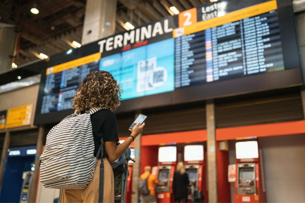 Eine Person mit lockigem Haar und gestreiftem Rucksack steht in einem Flughafenterminal vor einer großen Anzeigetafel für Abflüge und blickt auf ihr Handy.