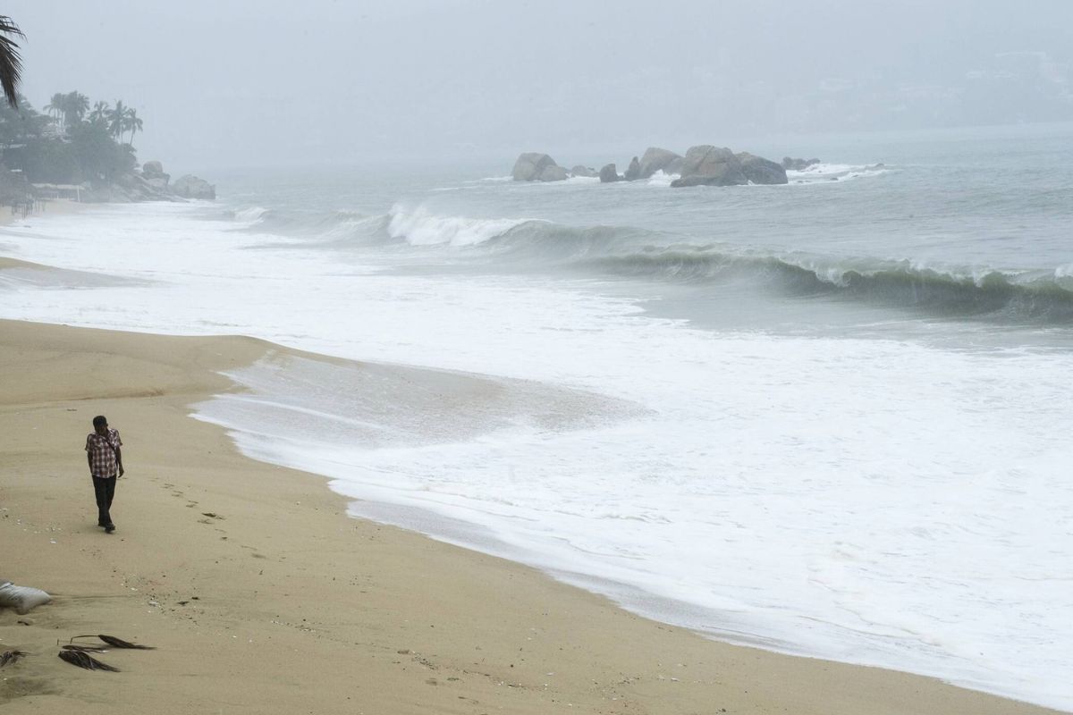 Ein einzelner Mensch geht bei bewölktem, stürmischem Wetter an einem Sandstrand entlang, während im Hintergrund Wellen auf Felsen im Meer treffen.