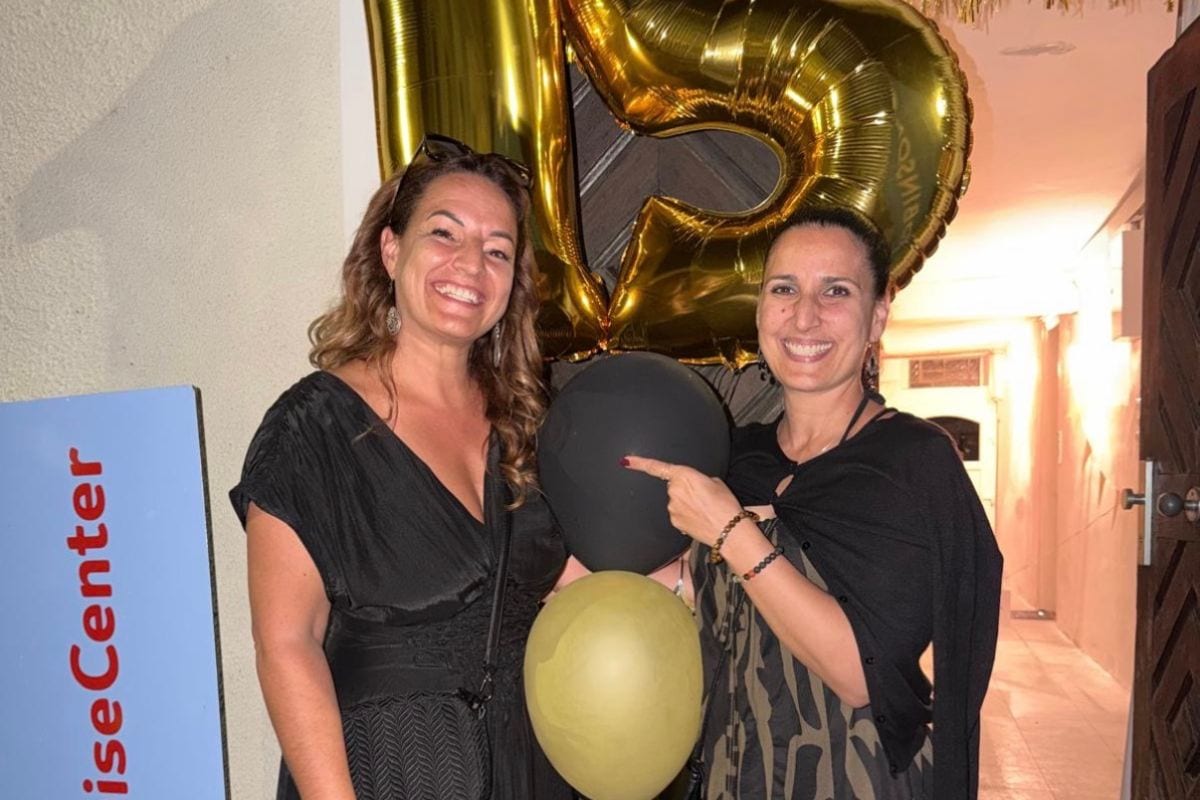 Zwei lächelnde Frauen stehen vor einer goldenen Ballon-Zahl 15, wobei eine Frau auf einen schwarzen Ballon zeigt, der über einem beigen Ballon schwebt.
