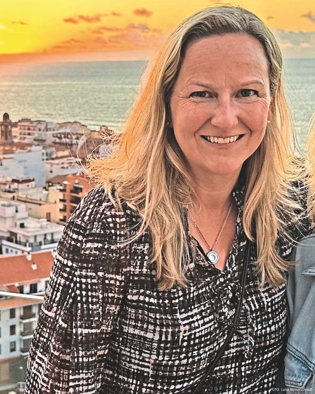 Eine blonde Frau mit schulterlangem Haar lächelt in die Kamera. Im Hintergrund ist eine Stadt am Meer während eines goldenen Sonnenuntergangs zu sehen.