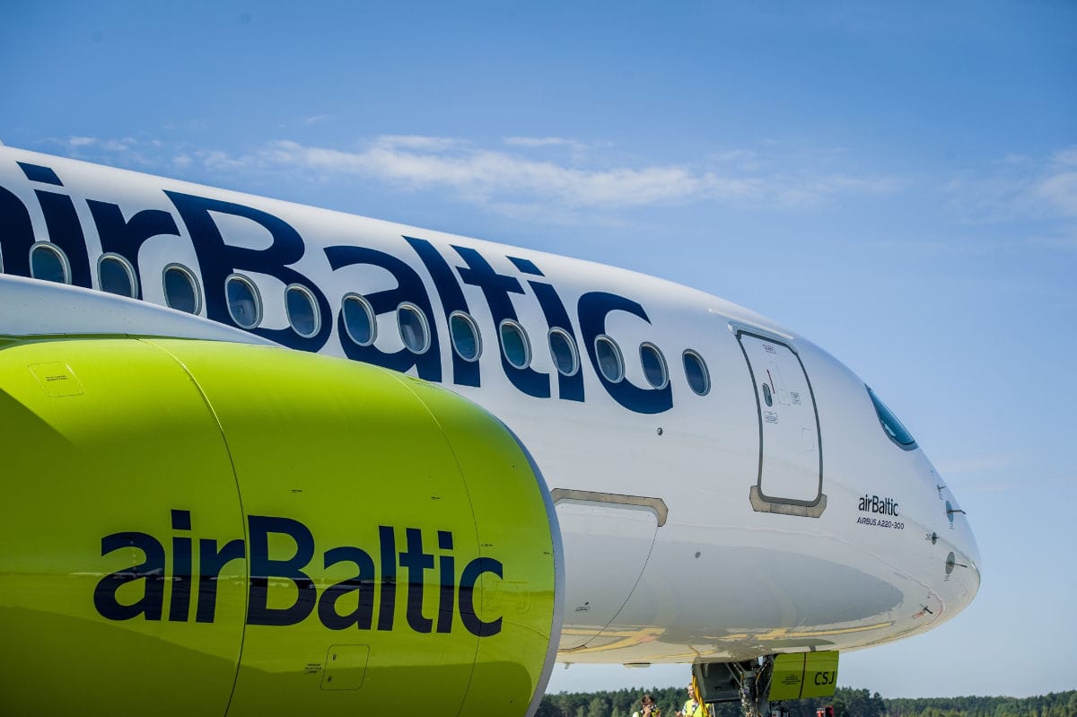 Nahaufnahme des vorderen Rumpfes und eines hellgrünen Triebwerks eines weißen airBaltic Airbus A220-300 Passagierflugzeugs vor einem klaren blauen Himmel.