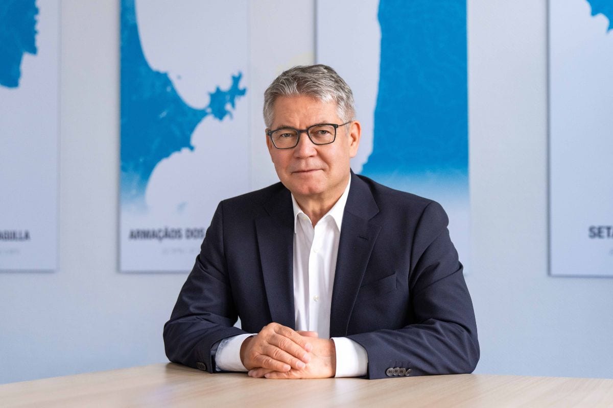 Ein Mann mit grauem Haar und Brille trägt ein weißes Hemd sowie ein dunkles Sakko und sitzt mit verschränkten Händen an einem Tisch vor blauen Grafiken.