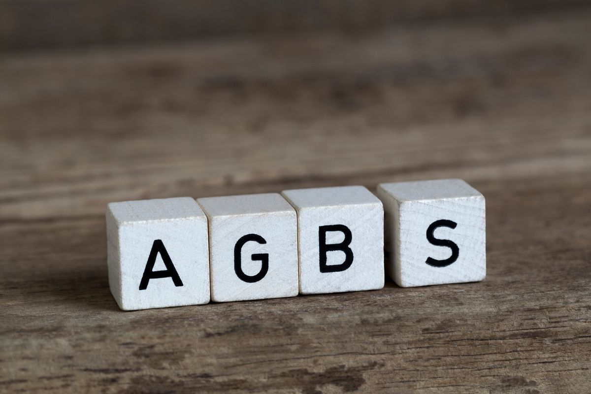 Vier weiße Holzwürfel mit den schwarzen Buchstaben A, G, B und S stehen in einer Reihe auf einem rustikalen, braunen Holzhintergrund mit sichtbarer Maserung.