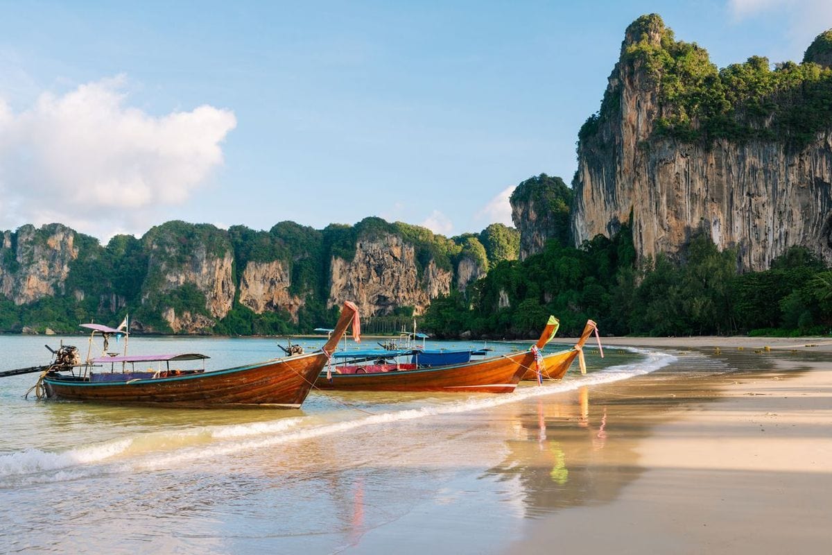 Mehrere traditionelle thailändische Langheckboote liegen am hellen Sandstrand vor einer Kulisse aus steilen, bewaldeten Kalksteinfelsen unter blauem Himmel.