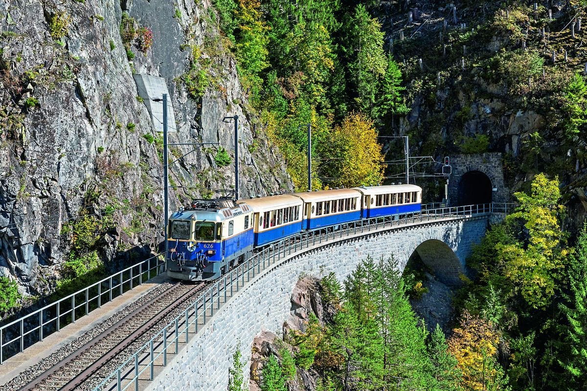 Ein blau-beiger Zug fährt auf einer steinernen Bogenbrücke vor einer steilen Felswand und einem Tunnelportal durch eine herbstliche Waldlandschaft.