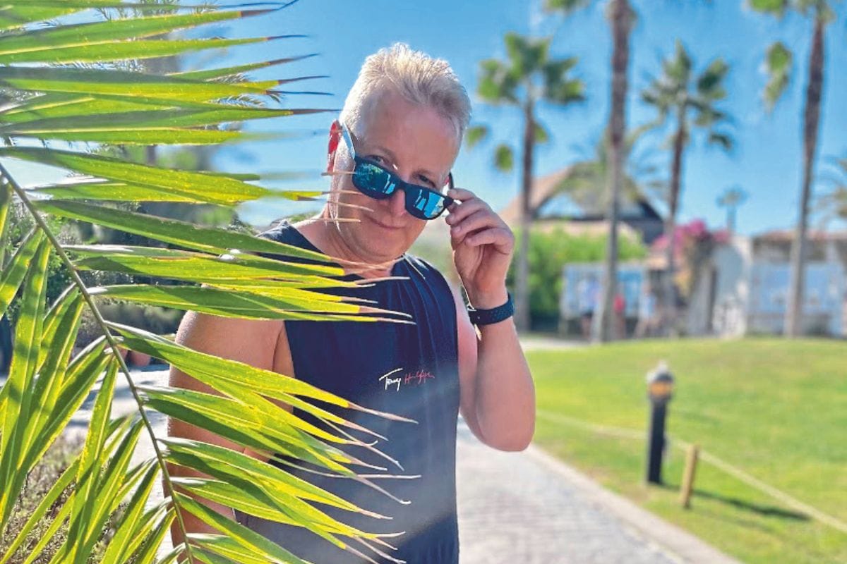 Ein Mann mit kurzem Haar und Sonnenbrille trägt ein dunkles ärmelloses Oberteil und steht vor einem Palmenblatt in einer sonnigen Hotelgartenanlage.