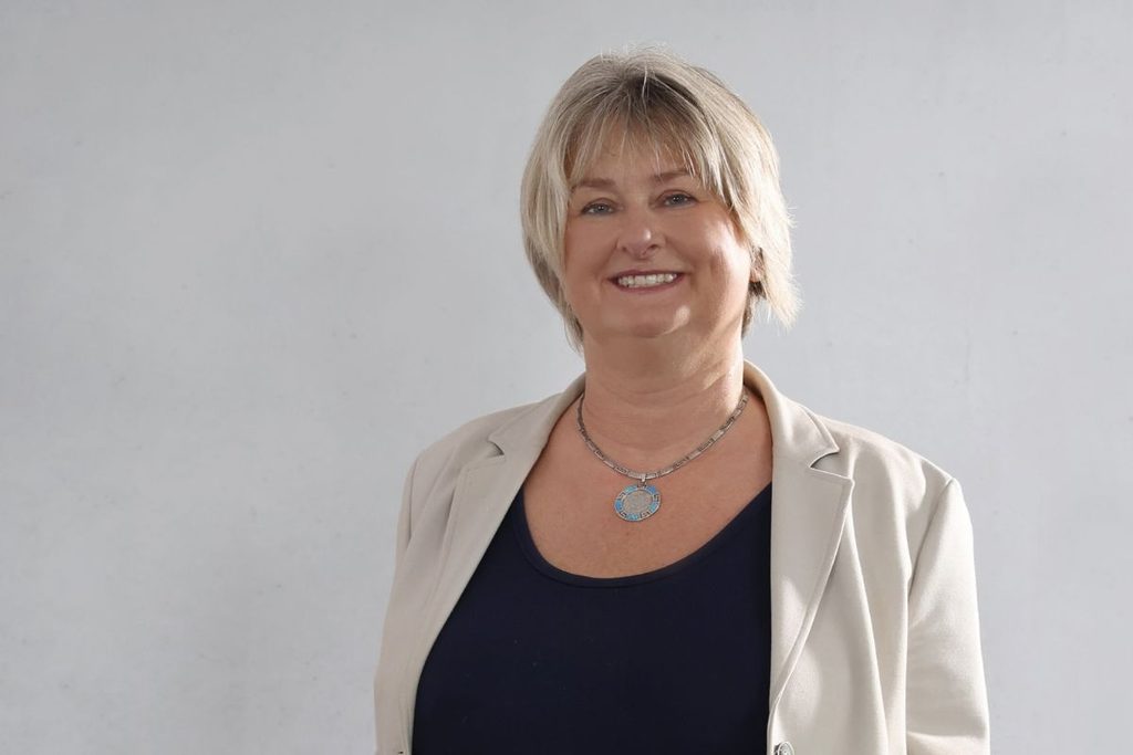 Eine Frau mit blondem Kurzhaarschnitt trägt ein dunkelblaues Oberteil unter einem hellen Blazer und eine silberne Halskette vor einem hellgrauen Hintergrund.